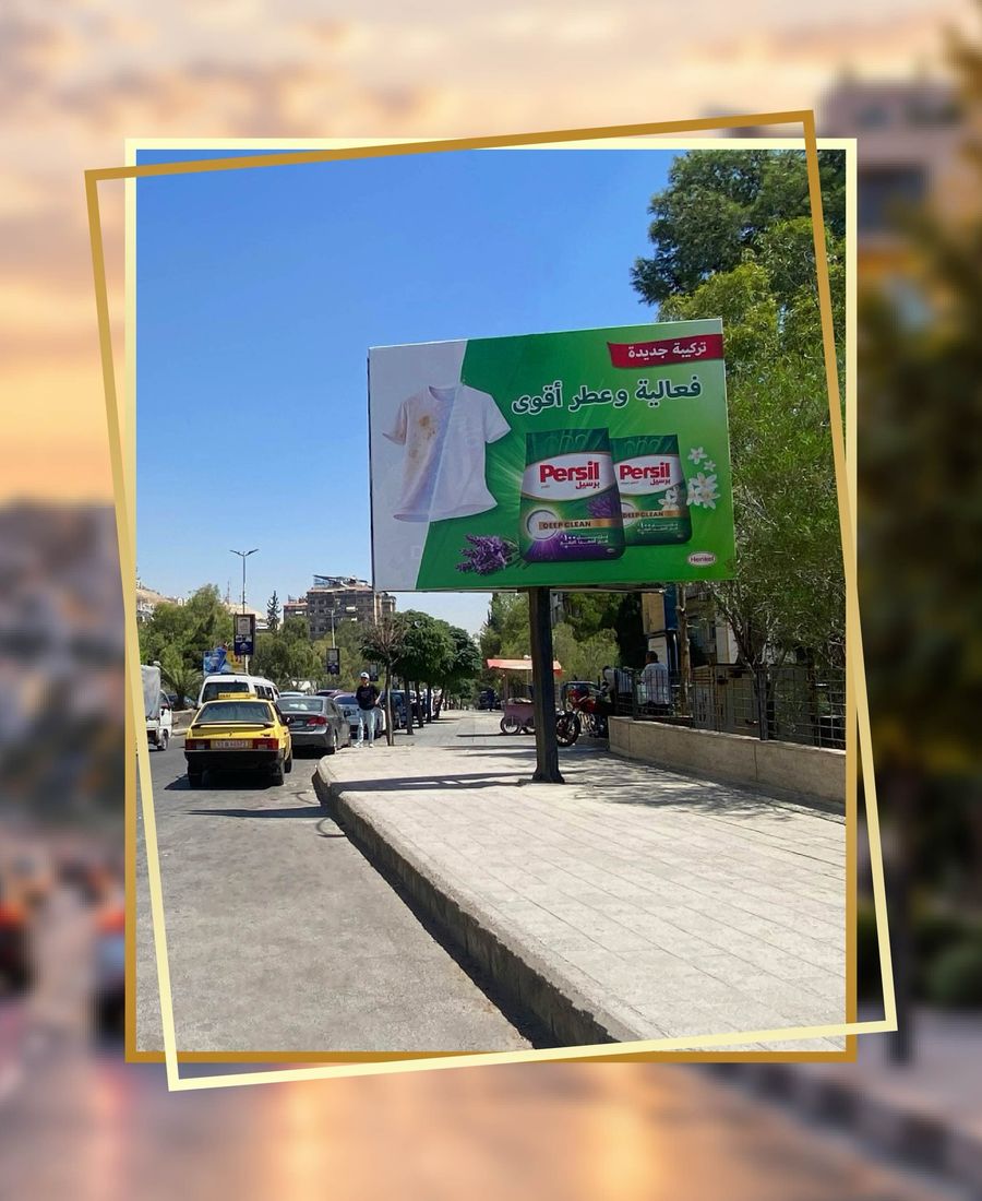 Persil billboard on a single column above a wide pavement, a yellow taxi and parked cars in the street