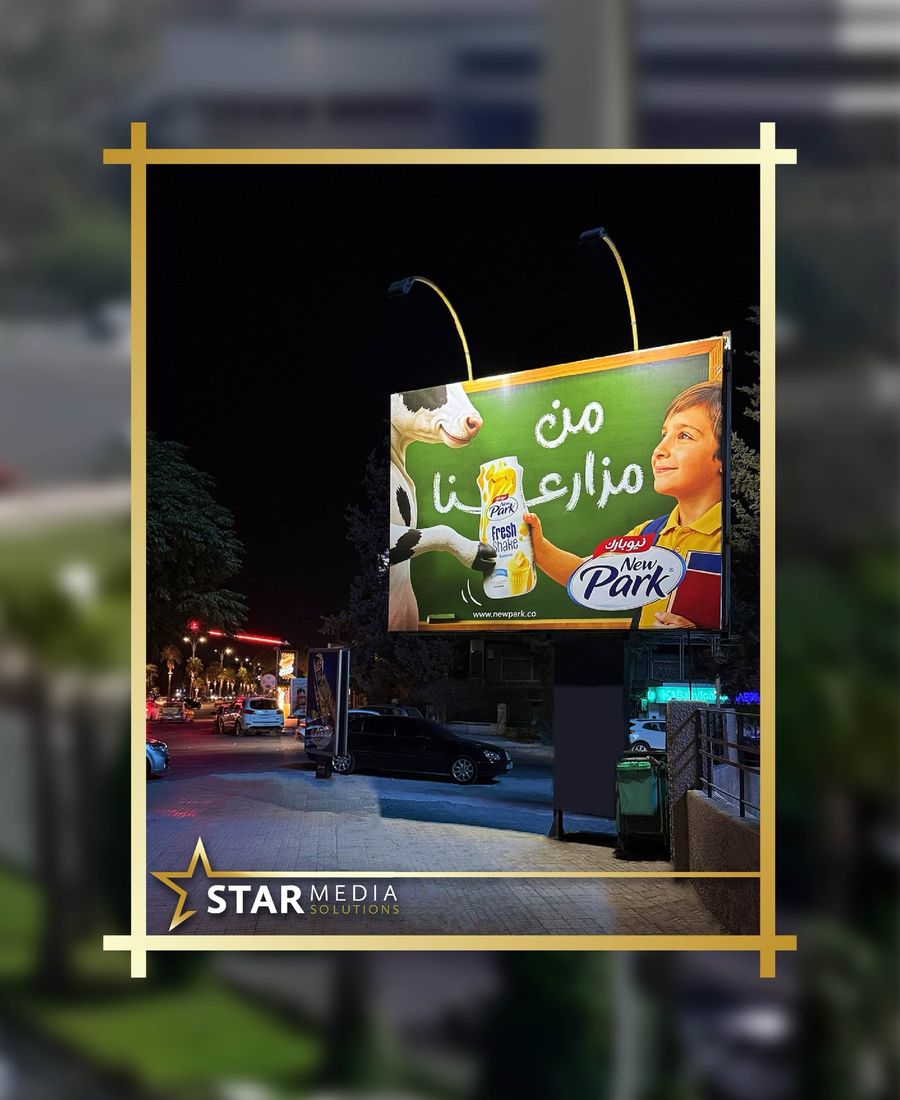 New Park billboard lit after dark on a Damascus street, two lamps angled down onto the face